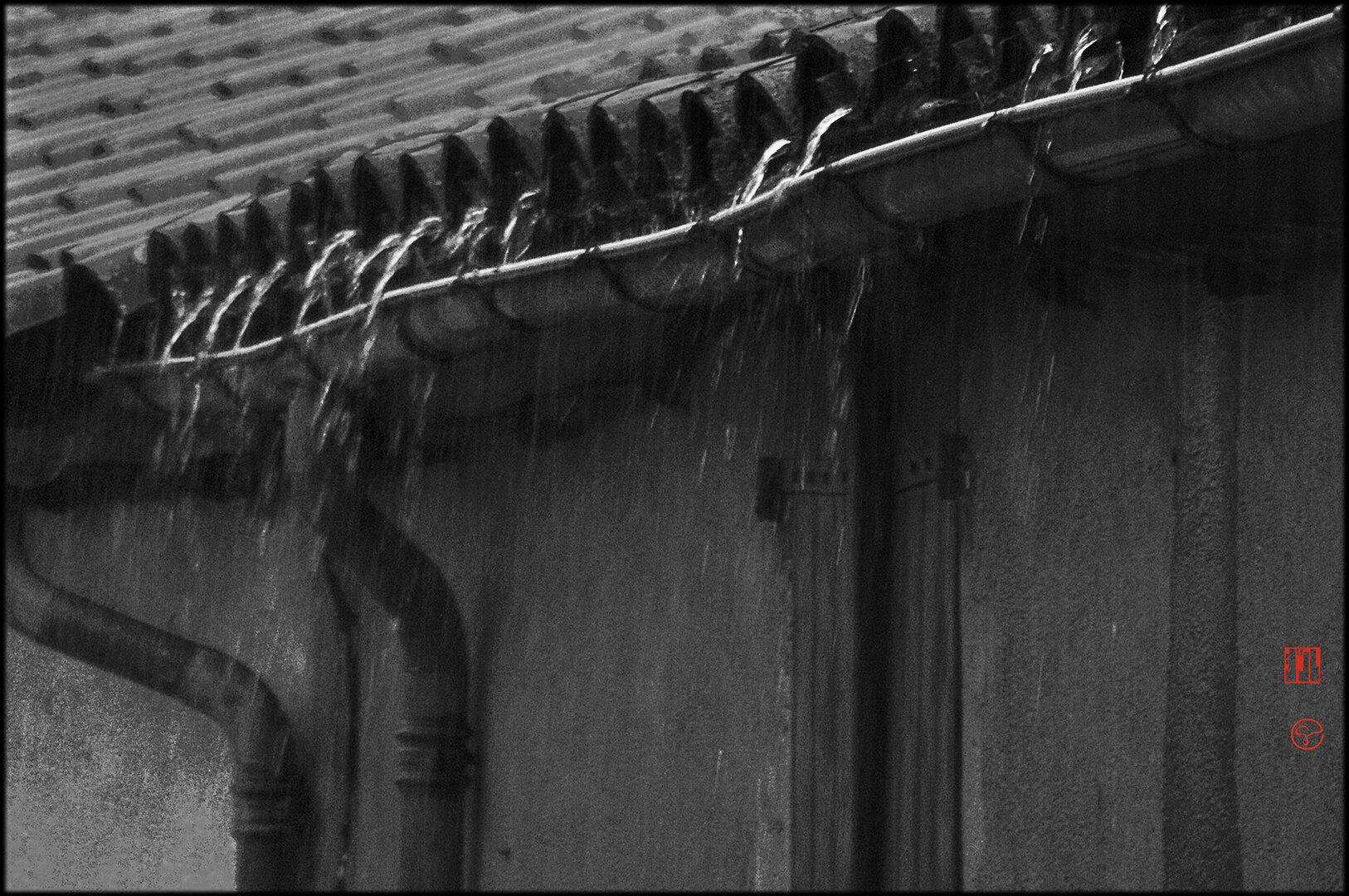 Format paysage, noir et blanc.
Dans une ambiance sombre, fortement contrastée, en diagonale descendant de l'angle droit vers environ le milieu de la marge gauche, sous un toit de tuiles rondes des gouttières en zinc affaissées ça et là. Une pluie intense fait gicler l'eau du toit au delà des gouttières et tombe directement en cascades dans le vide